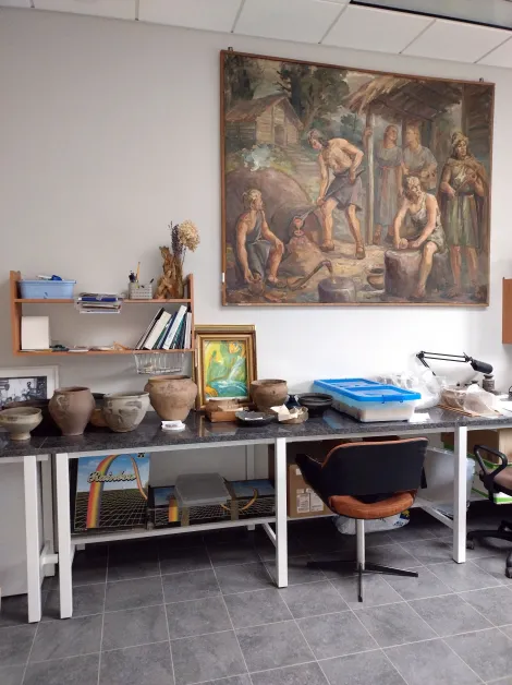 Pomieszczenie dokumentacyjne PKC. Na stole ustawione są zrekonstruowane naczynia w trakcie wysychania i dokumentowania.  alt: Długi stół pod ścianą. Na stole zrekonstruowane gliniane naczynia. Naczynia w odcieniach brązu i szarości mają różne rozmiary.