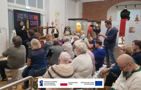 tłum osób zgromadzonych na sali wystawowej oraz prowadząca wykład prowadząca ustawiona frontem do publiczności. Za nią banner promujący wystawę, w tle gabloty z manekinami oraz zabytkami