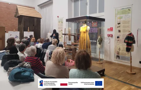 grupa osób siedzących na krzesłach i słuchających wykładu, po ich lewej stronie duży drewniany młyn wietrzny, po prawej gabloty