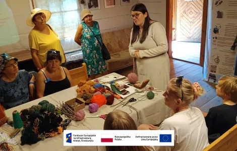 grupa kobiet zebrana przy stole warsztatowym. Między nimi stojąca prowadząca warsztaty, w okularach i z długimi rozpuszczonymi włosami, ubrana w lnianą długą sukienkę