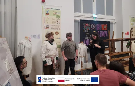 po lewej: mężczyzna w średnim wieku, z okazałą brodą oraz w okularach, ubrany w średniowieczny strój (lnianą koszulę, czapę z futrem oraz sznurkowy pasek), po prawej drugi mężczyzna w okularach oraz prowadząca wykład - kobieta z długimi ciemnymi włosami oraz w okularach