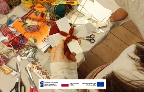 na drewnianym stole przykrytym częściowo lnianym płótnem leżą: książka z pomarańczową okładką, motki kolorowej włóczki, ścinki materiałów i nożyczki, na pierwszym planie zbliżenie na tworzony przez uczestniczkę warsztatów kwiat z czerwonej włóczki 