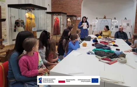 duży stół nakryty białą tkaniną, na którym znajdują się kolorowe włóczki oraz inne materiały potrzebne do warsztatów. Przy stole siedzą dzieci i dorośli i słuchają prowadzącej warsztaty