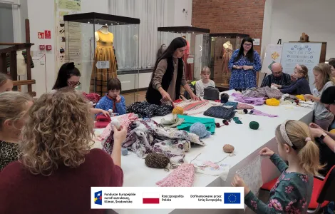 kobieta z długimi, prostymi ciemnymi włosami rozkładająca duży kawałek czerwono - białego materiału na stole. Wokół niej siedzące przy stole uczestniczki warsztatów 