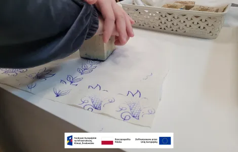 dwie dłonie dociskające drewniany stempel, przy pomocy którego odbijany jest na materiale kwiatowy wzór