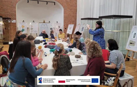 grupa kobiet z dziećmi oraz jeden mężczyzna siedzący przy stole warsztatowym, na którym znajdują się kolorowe materiały