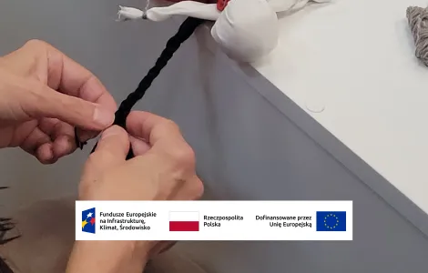 leżąca na stole lalka motanka w czerwonej spódniczce z białym fartuszkiem. Dwie dłonie zaplatają jej na głowie brązowe warkocze 