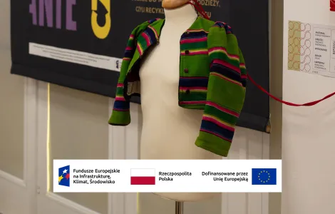 Manekin ubrany w krótki, zielony żakiet w kolorowe, poziome pasy stoi na tle czarnego plakatu wystawy z dużym napisem „CEROWANIE CZASU”. Ekspozycja znajduje się w jasnym wnętrzu, a na dole widoczne są logotypy Funduszy Europejskich, Rzeczypospolitej Polskiej i Unii Europejskiej.