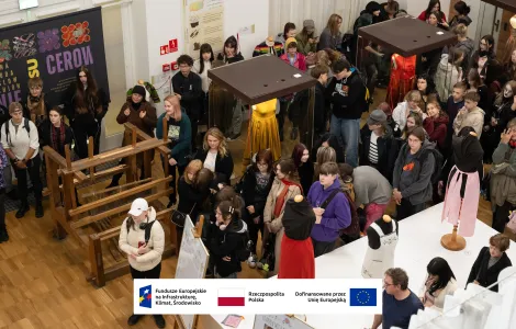 Grupa kilkudziesięciu osób zwiedza wystawę w sali muzealnej. W centrum stoi duże drewniane krosno tkackie, obok gabloty z manekinami w kolorowych sukniach (m.in. żółtej i czerwonej). Zwiedzający stoją blisko siebie i oglądają eksponaty. Widoczny jest plakat wystawy oraz logotypy Funduszy Europejskich i Unii Europejskiej.