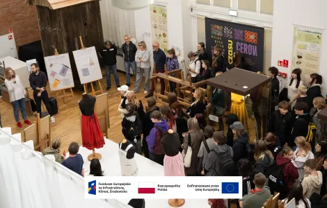 Widok z góry na salę wystawową wypełnioną zwiedzającymi. Kobieta z mikrofonem przemawia do grupy młodzieży i dorosłych stojących wokół ekspozycji. W centrum znajdują się manekiny w kolorowych strojach (m.in. czerwonej i różowej spódnicy), drewniane krosno tkackie oraz plansze edukacyjne. Na ścianie widać duży plakat z napisem „CEROWANIE CZASU”, a na dole grafiki – logotypy Funduszy Europejskich, Rzeczypospolitej Polskiej i Unii Europejskiej.