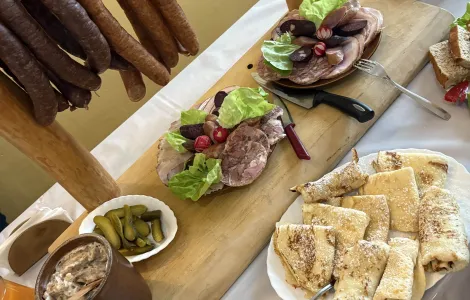 zastawiony stół z poczęstunkiem, na którym znajdują się pęta kiełbasy, ogórki kiszone, smalec w glinianym garnku, naleśniki oraz półmiski w wędlinami