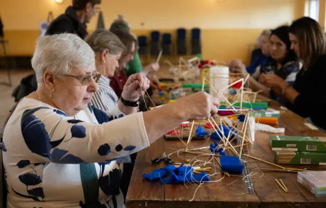 zdjęcie przedstawiające grupę ludzi siedzących przy stole podczas warsztatów z tworzenia słomkowych pająków. Na pierwszym planie starsza kobieta w okularach nawlekająca na nitkę kawałek słomy. 