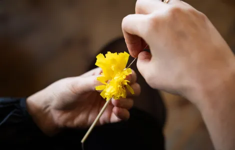 zbliżenie na dłonie, które przewlekają przez nitkę kawałek żółtej bibuły, który stanowi ozdobę słomkowego pająka