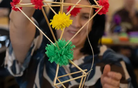 kobieta w średnim wieku z ciemnymi włosami trzymająca przed sobą słomkowego pająka z zielonymi, czerwony i żółtymi ozdobami z bibuły 