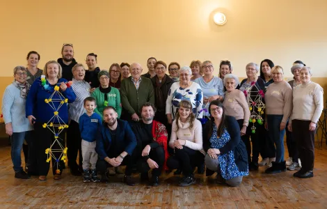 grupowe zdjęcie uczestników warsztatów, po lewej i po prawej stronie kobiety trzymające powstałe podczas warsztatów słomkowe pająki 