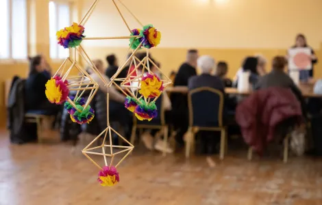 słomkowy pająk z kolorowymi ozdobami z bibuły, w tle uczestnicy warsztatów siedzący przy stole 