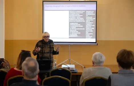 starsza kobieta z jasną grzywką i okularami w grubych oprawkach, ubrana w brązową sukienkę, podczas wystąpienia. Za nią na ścianie fragment prezentacji z napisem "regiony etnograficzne w województwie łódzkim" 