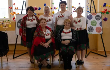 6 kobiet z koła gospodyń wiejskich, ubranych w jednakowe białe koszulki z kwiatowym wzorem i czerwone korale. Dwie z nich na plecach mają czerwoną wzorzystą chustę. W tle plakaty na których wydrukowane są wycinanki