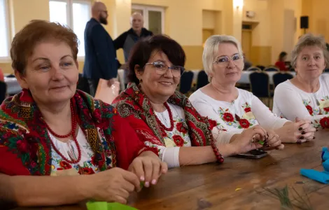 4 kobiety siedzące przy stole, dwie z nich mają czerwone korale na szyi oraz czerwone chusty na ramionach, dwie kolejne ubrane w jednakowe białe koszulki z kwiatowym motywem 