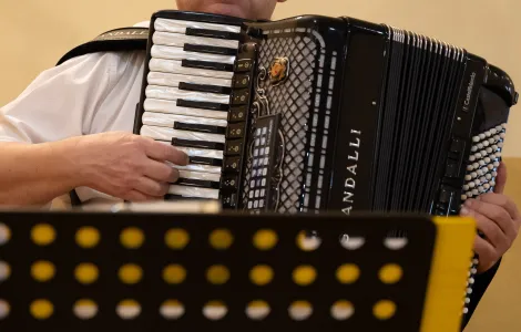 starszy mężczyzna w kaszkiecie i okularach z harmonią/ akordeonem. Przed nim pulpit z nutami 