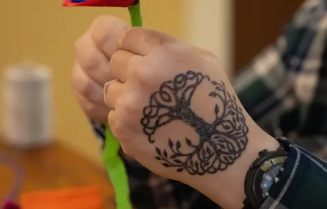 zbliżenie na dłonie (na jednej z nich tatuaż przedstawiający drzewo), które trzymają tworzony kwiat z niebieskiej, czerwonej i zielonej bibuły 