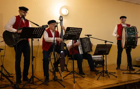 Kapela Łęczycka, od lewej wysoki mężczyzna z gitarą, mężczyzna grający na elektrycznym kontrabasie, siedzący mężczyzna z harmonią oraz wysoki mężczyzna grający na bębnie. Wszyscy ubrani w jednakowe stroje: białe koszule, pasiaste pomarańczowe kamizelki, czarne spodnie oraz kaszkiety na głowach