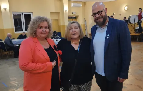 kobieta z kręconymi włosami ubrana w koralową marynarkę i ciemną sukienkę, kobieta w ciemnej marynarce oraz wysoki mężczyzna z brodą i w okularach.