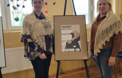 dwie kobiety w chustach kamelowych stojące przy sztaludze z plakatem