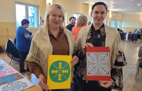 kobieta z jasnymi włosami i zarzuconą na plecy chustą kamelową oraz kobieta z ciemnymi włosami również w kamelowej chuście, trzymają przygotowane wycinanki. 