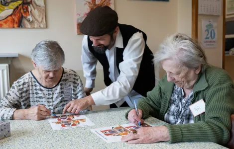 Dwie starsze kobiety siedzą przy stole. Między nimi stoi mężczyzna w białej koszuli i czarnej czapce. Kobiety zaznaczają długopisami coś na kolorowych kartkach.
