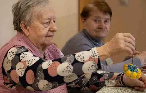 Dwie starsze kobiety siedzą przy stole. Kobieta na pierwszym planie trzyma w ręku zółto - niebieską piłeczkę na gumce.