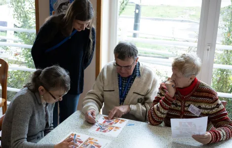Przy stole siedzą trzy osoby. Między nimi stoi młoda kobieta. Ludzie patrzą na kartki z kolorowymi obrazkami, trzymają w rękach długopisy.