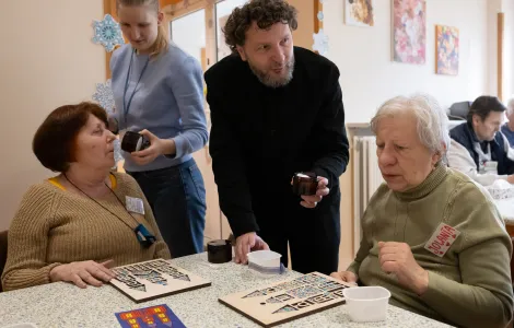 Dwie starsze kobiety siedzą przy stole. Przed nimi leżą drewniane szablony szopek krakowskich. Obok nich stoi mężczyzna i kobieta, którzy trzymają w ręku słoiczki. Jedna ze starszych kobiet wącha słoiczek.