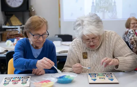 Dwie starsze panie siedzą przy szarym stole. Przed nimi leżą drewniane szablony przedstawiające budynek. Wsypują łyżeczkami kolorowy ryż do okienek tego budynku.