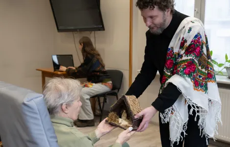 Mężczyzna w czarnej bluzce i chustce w kwiaty na ramieniu pokazuje starszej pani szopkę betlejemską. Razem trzymają tę rzeźbę w rękach.