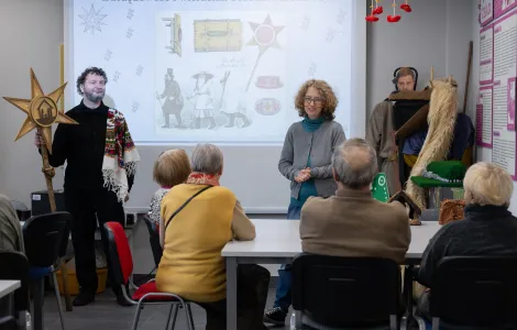 Na tle ekranu z obrazkiem i  z napisem "Obrzędowość i wierzenia bożonarodzeniowe" stoją kobieta i mężczyzna. Mężczyzna trzyma gwiazdę kolędniczą. Grupa osób siedzi przy stoliku twarzą do ekranu.