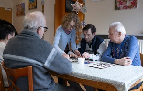 Grupa starszych mężczyzn siedzi przy stole. Na stole leżą drewniane szablony szopek krakowskich. Obok jednego z mężczyzn stoi kobieta. Razem robią coś przy szablonie.