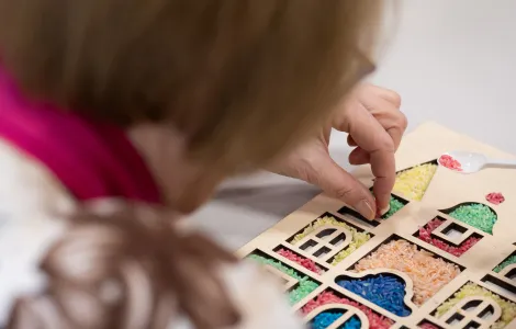 Jakaś osoba wrzuca ziarenka ryżu do drewnianego szablonu. W szablonie są okienka o różnych kształtach pełne kolorowego ryżu.