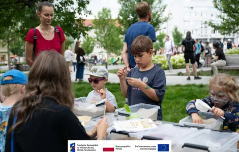 Grupa dzieci podczas warsztatów archeologicznych przy stole piknikowym rozstawionym na zielonej trawie na placu Wolności. W tle tłum osób zebranych na placu. 