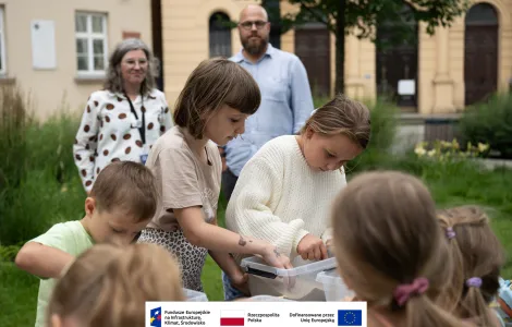 Na pierwszym planie grupa dzieci podczas warsztatów archeologicznych. Na stole stoi plastikowy pojemnik wypełniony piaskiem, wśród którego dzieci próbują odnaleźć ukryte skarby. W tle budynek muzeum oraz kobieta z szarymi włosami w okularach oraz wysoki mężczyzna z brodą w błękitnej koszuli
