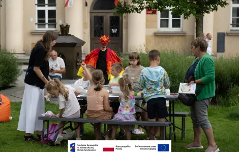 Grupa dzieci podczas warsztatów archeologicznych siedząca przy stole piknikowym. Po lewej animatorka nadzorująca zajęcia, po prawej kobieta w średnim wieku. W tle fontanna miłości z placu Wolności, mężczyzna w średnim wieku, mężczyzna w stroju feniksa oraz budynek muzeum. 