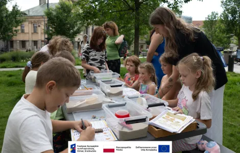 Grupa osób podczas warsztatów archeologicznych siedząca przy stole piknikowym rozstawionym na zielonej trawie na placu Wolności. W tle budynek archiwum oraz przypadkowi przechodnie. 