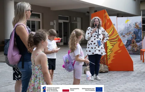 Po prawej stronie kobieta z szarymi włosami w okularach, ubrana w jasną koszulę w brązowe wzory przemawia do mikrofonu podczas otwarcia wystawy. Na prawo od kobiety widoczna rozstawiony fragment mobilnej wystawy plenerowej, która wykonana jest z połączonych ze sobą pionowych elementów (ścianek) utrzymanych w pomarańczowej kolorystyce. Po lewej stronie goście zaproszeni na otwarcie wystawy. W tle budynek na dziedzińcu muzealnym.