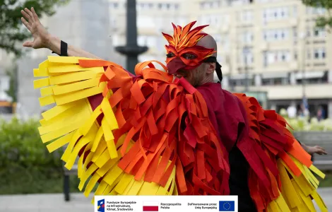 Uśmiechnięty mężczyzna ubrany w strój mitycznego ptaka – feniksa. Na twarzy ma maskę imitującą dziób ptaka, na ramionach pelerynę imitującą pióra ptaka w kolorach intensywnej czerwieni i odcieniach żółtego. Mężczyzna ma rozłożone ręce udając, że rozkłada „skrzydła”. 