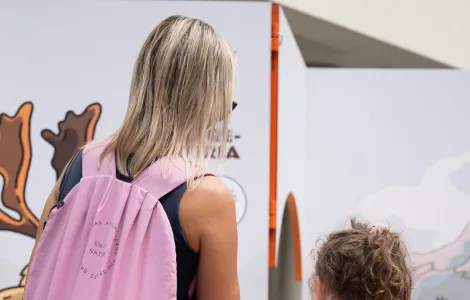Na pierwszym planie młoda kobieta z dzieckiem stojące tyłem od obiektywu. Kobieta ma jasne włosy oraz różowy plecak, dziecko jest ubrane w kwiecistą letnią sukienkę. Oby dwie patrzą na jedną z ekspozycji na ściance wystawowej. 