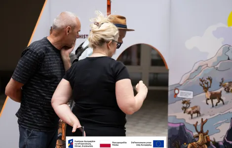 Grupa osób – kobieta w średnim wieku uczesana kok oraz dwóch mężczyzn w średnim wieku – jeden w kapeluszu słomkowym czytający napisy na jednej ze ścianek wystawowych. 