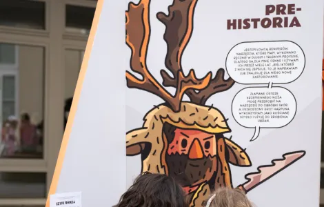 Jedna ze ścianek wystawowych zatytułowana „Prahistoria” przed którą kuca ubrana na ciemno kobieta z lekko kręconymi ciemnymi włosami wraz z małą dziewczynką w różowych getrach i wzorzystej tunice. Obie próbują rozwiązać quiz, który jest jednym z zadań przygotowanych dla uczestników wydarzenia. 