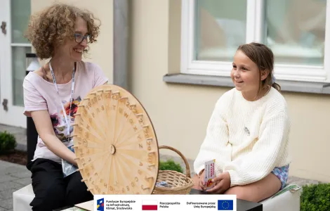 Kobieta w średnim wieku w okularach z krótkimi kręconymi włosami uśmiechająca się do dziewczynki siedzącej po jej prawej. Dziewczynka ma ciemne włosy i ubrana jest w ażurowy jasny sweter. Na pierwszym planie wykonane z drewna koło z literami służące do quizu etnograficznego. 