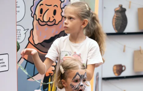 Dwie dziewczynki – jedna wyraźnie starsza, uczesana w kucyk i ubrana w różową koszulkę, druga młodsza we wzorzystej tunice oraz pomalowaną we wzór kotka twarzą podczas rozwiązywania „szyfru feniksa” na wystawie mobilnej 