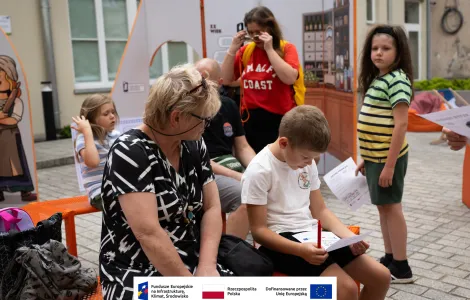 Grupa osób podczas otwarcia wystawy. Na pierwszym planie starsza kobieta w okularach i czarno-białej sukience wraz z chłopcem czytającym coś na trzymanej kartce. W tle inni zwiedzający wraz z dziećmi. 